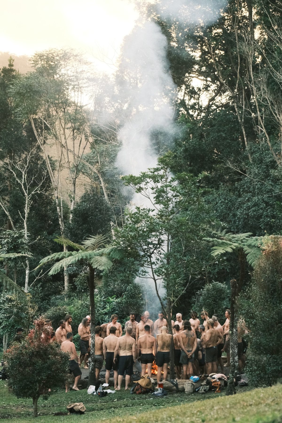 A large circle of men standing around a smoking fire in a rainforest clearing