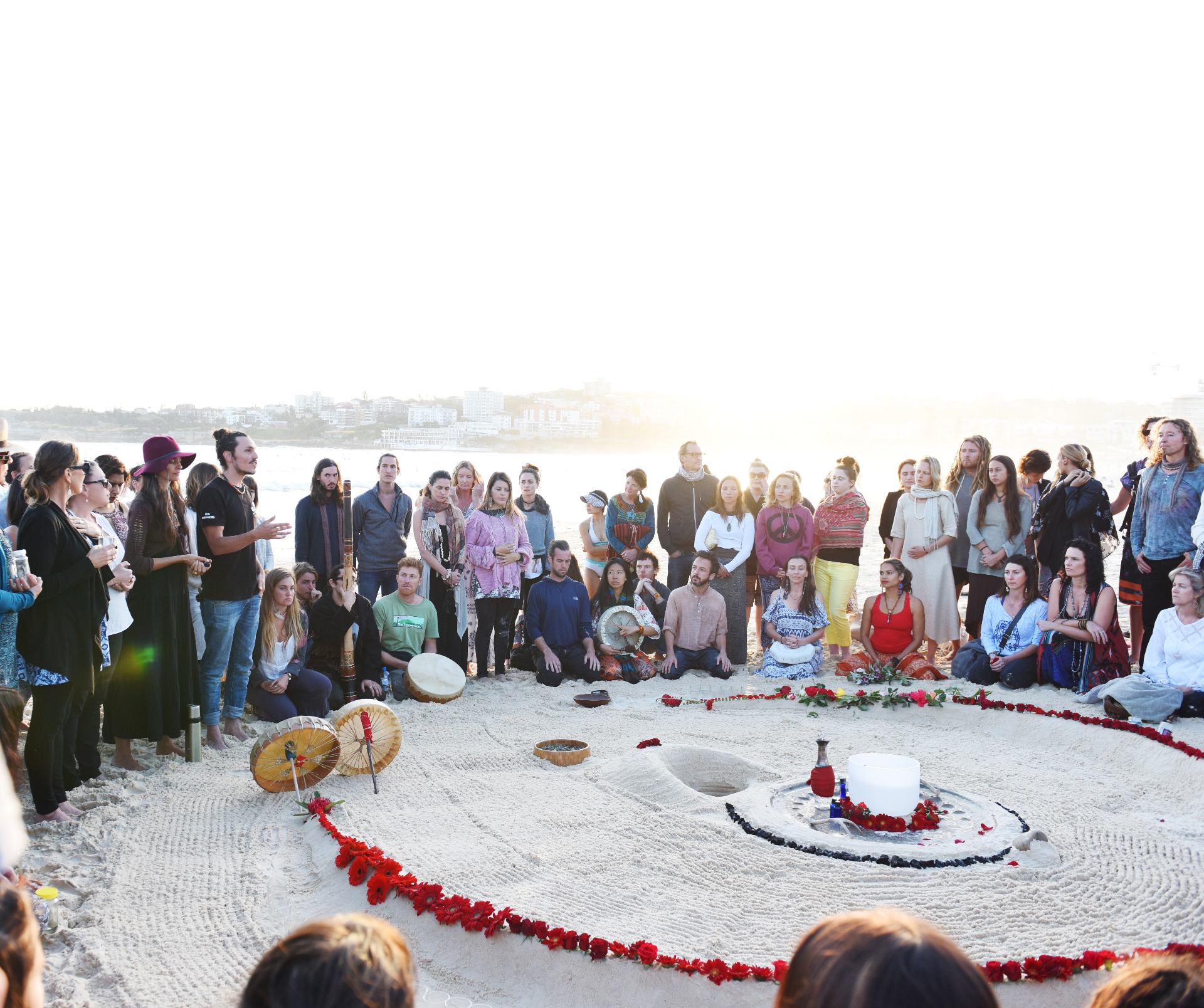 A large circle gathered at dawn around a ceremonial sand mandala ringed with red flowers on the beach