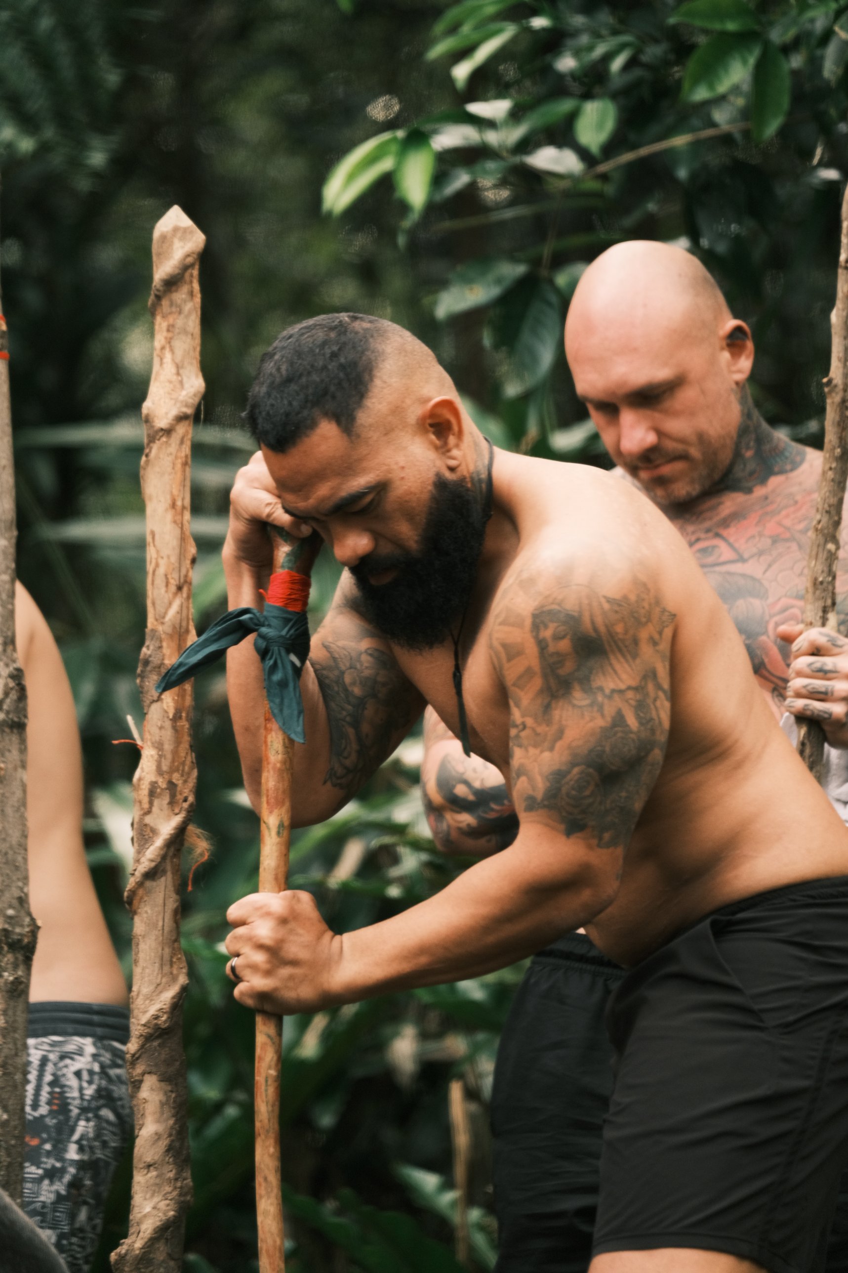 A bearded, tattooed man leans his head against a wrapped wooden staff in the rainforest, another man with a staff behind him