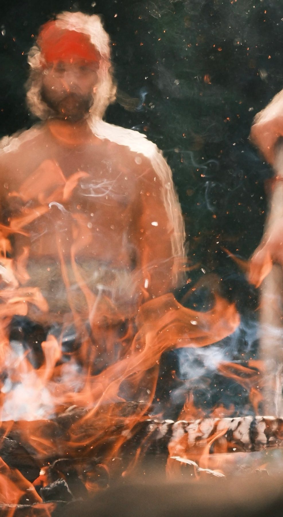 A man stands bare-chested behind a ceremonial fire, sparks and smoke rising between him and the camera