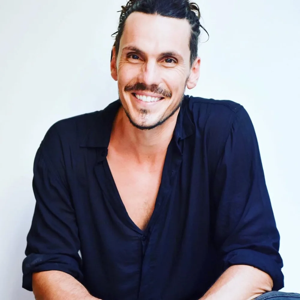 Portrait of Velan smiling, in a dark open-collared shirt against a pale wall