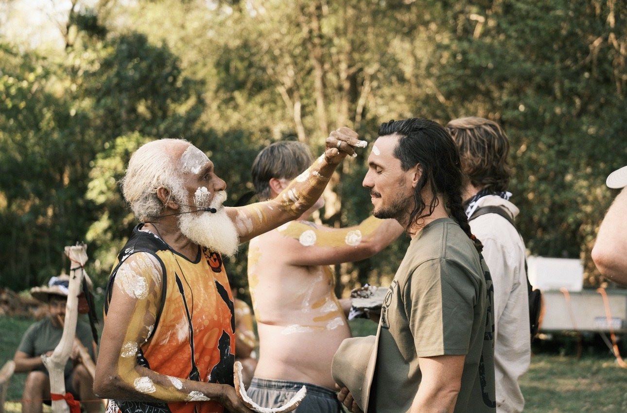 An Elder marks a participant's brow with white ochre during ceremony, other men painted and waiting behind