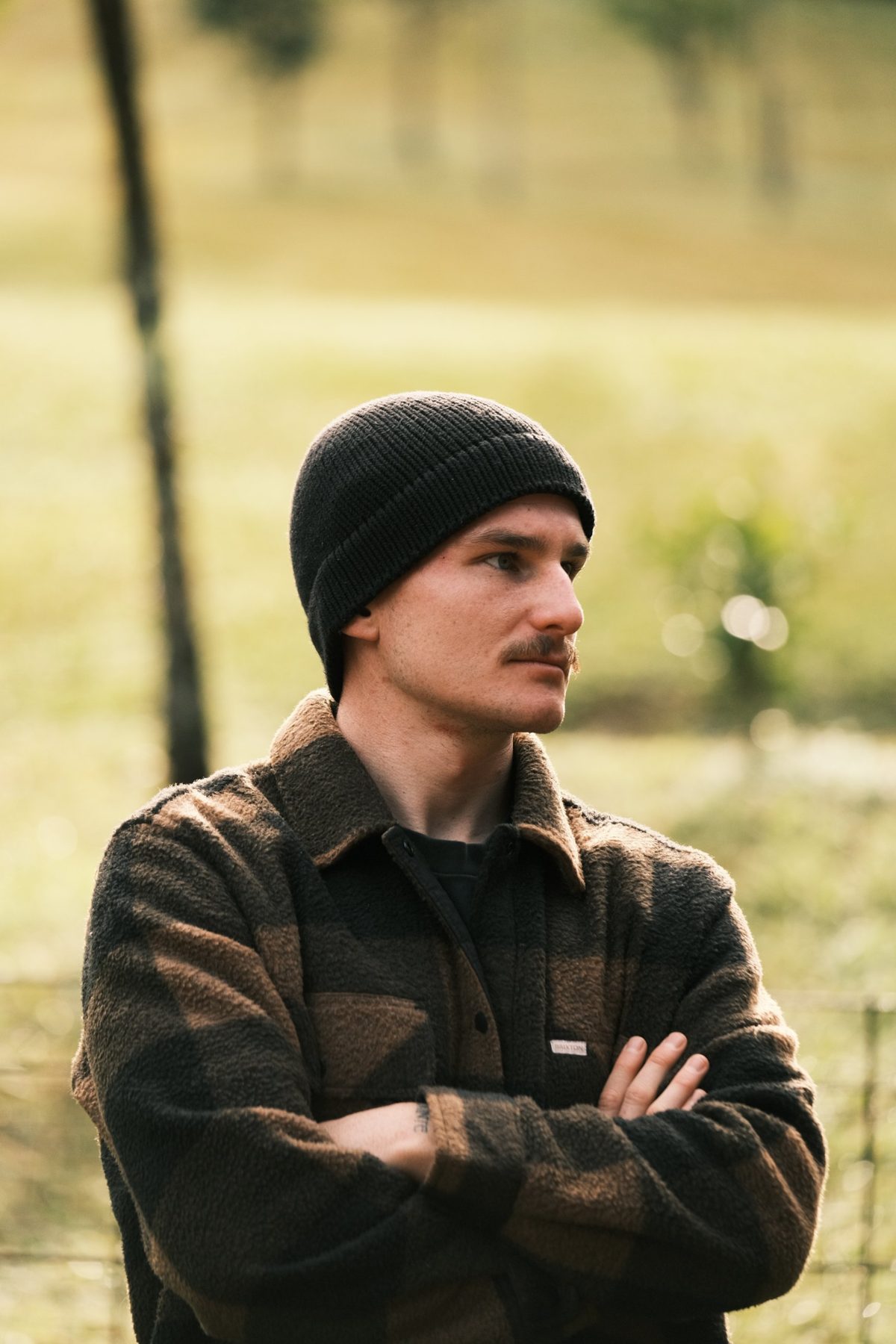 A man in a beanie and flannel jacket stands with arms crossed, looking out over the paddock